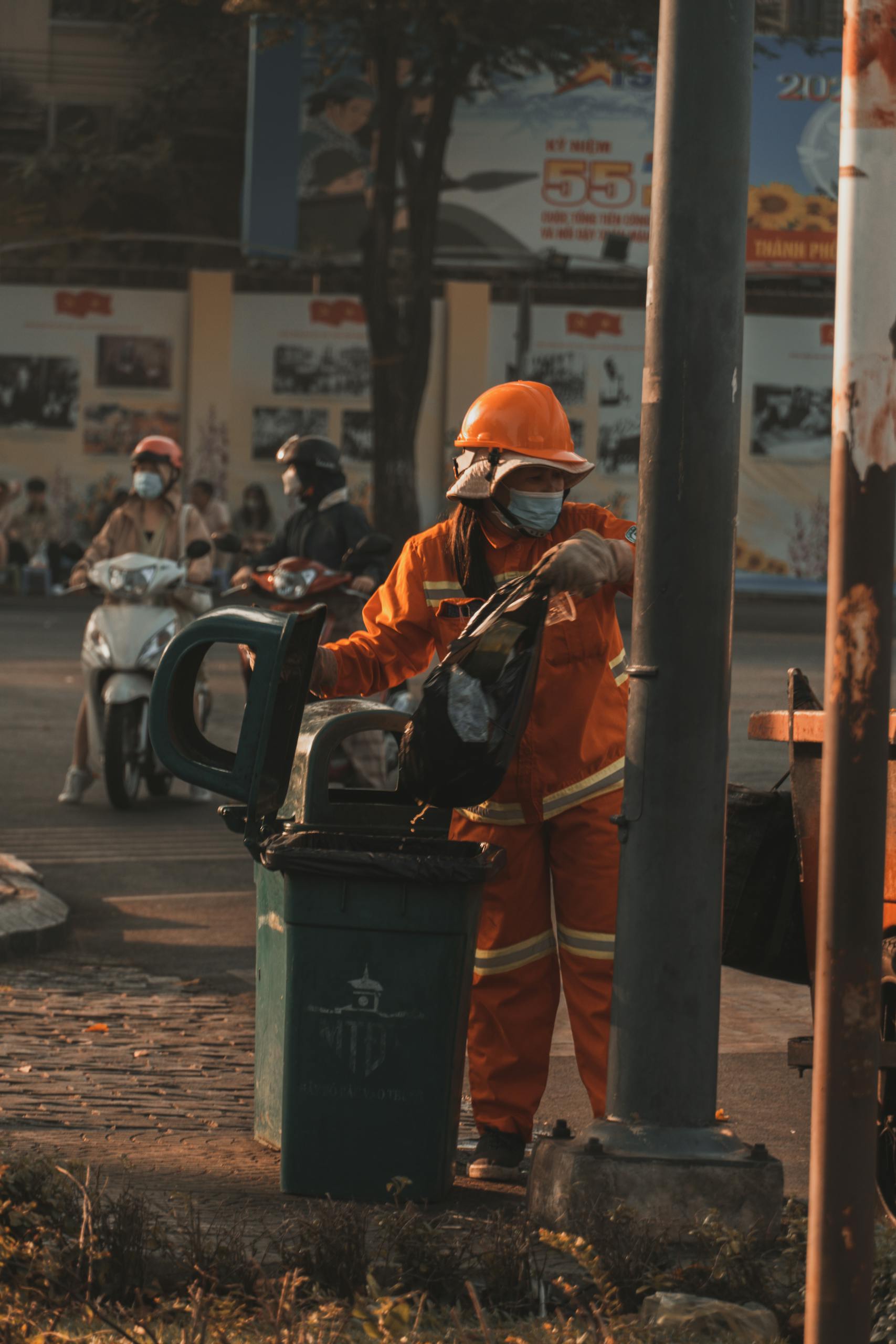 A sanitation worker in an orange suit empties a trash can on a city street.