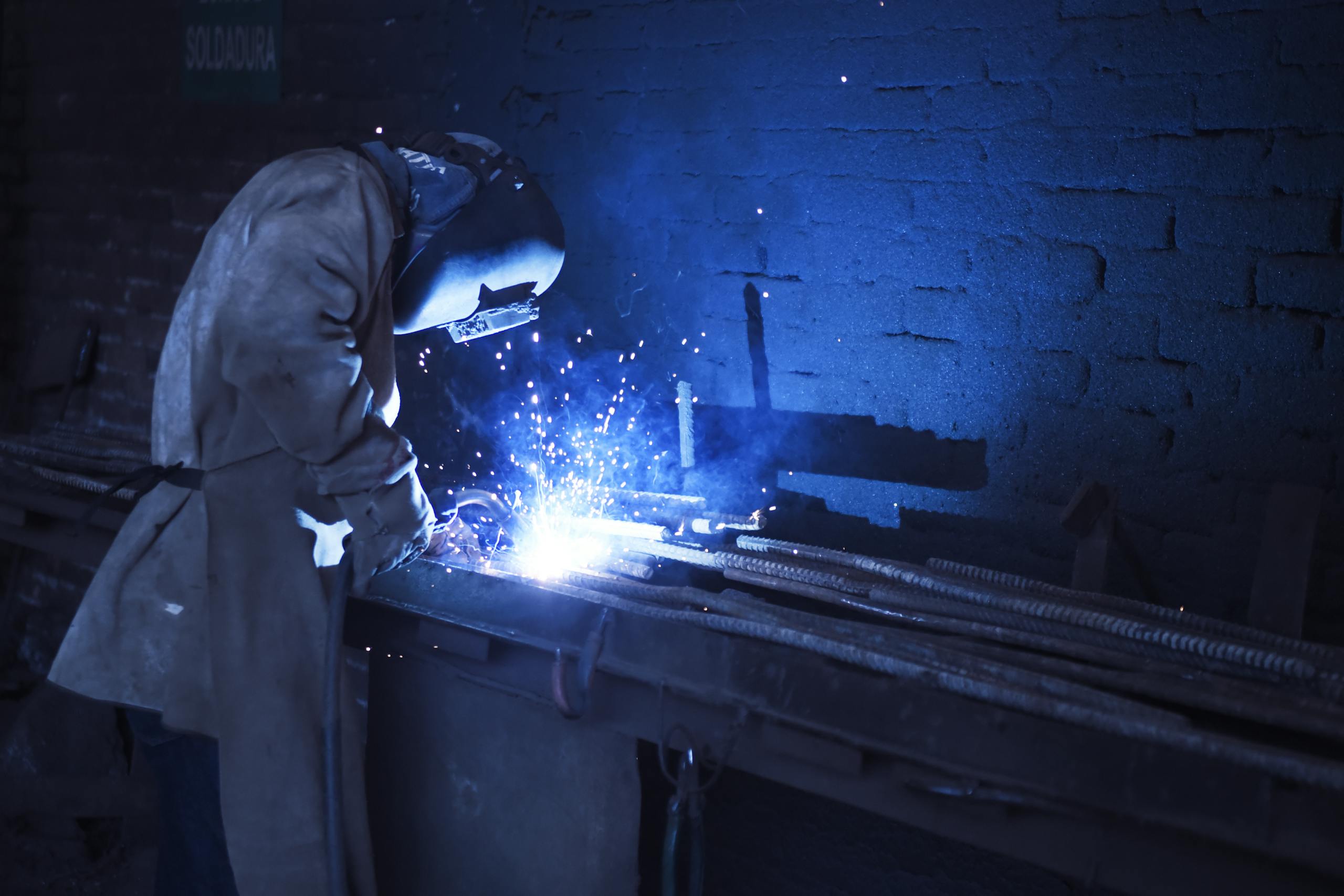 Industrial worker welding metal indoors, emitting bright blue sparks and smoke.